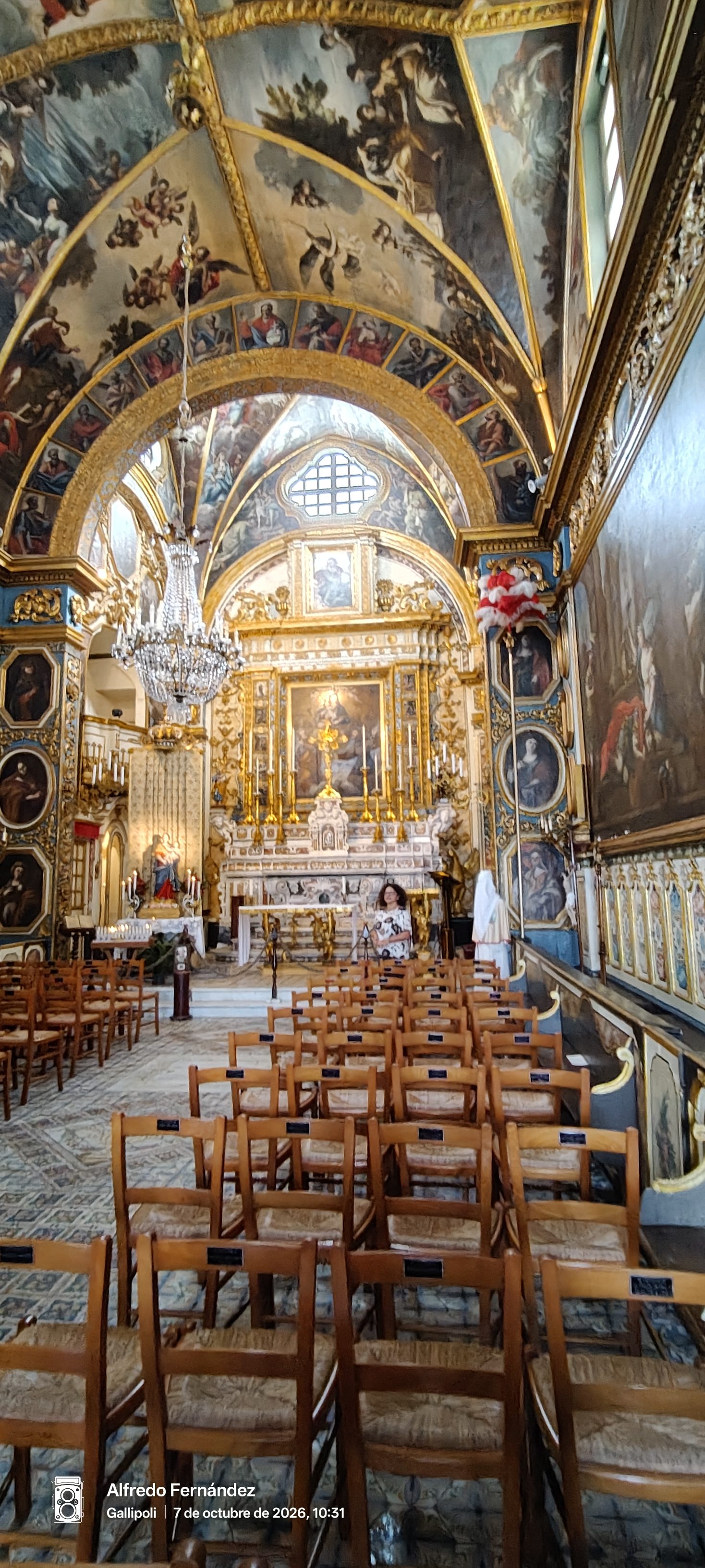 Interior barroco de iglesia con techos pintados al óleo, altar dorado y sillas de madera.