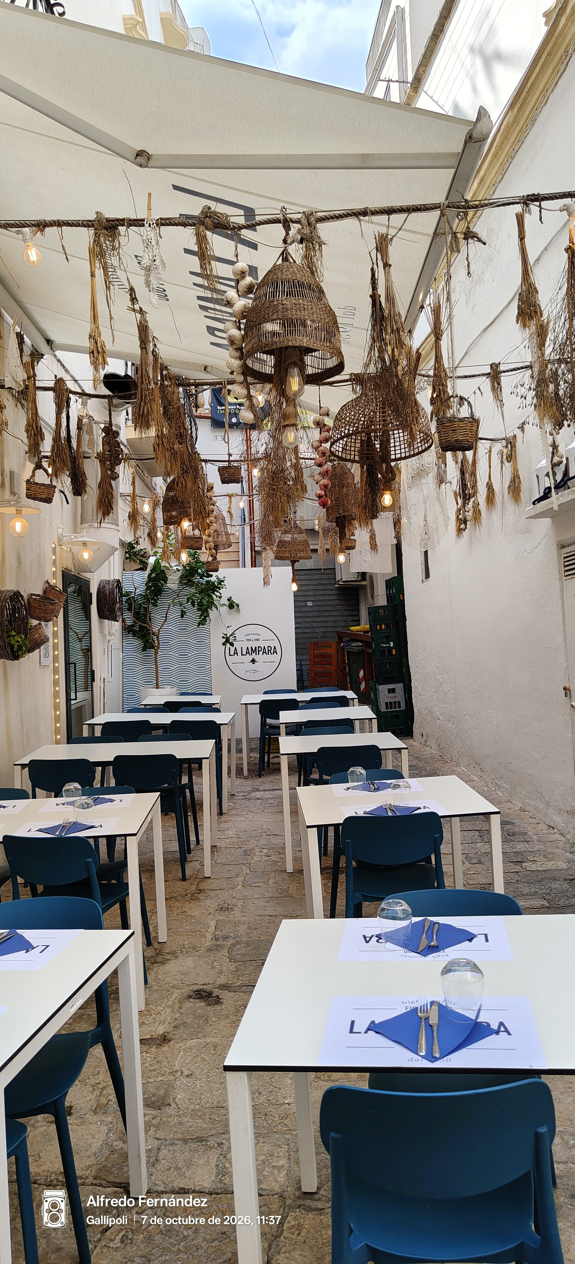 Mesas preparadas con sillas azules en un callejón decorado con lámparas de mimbre.