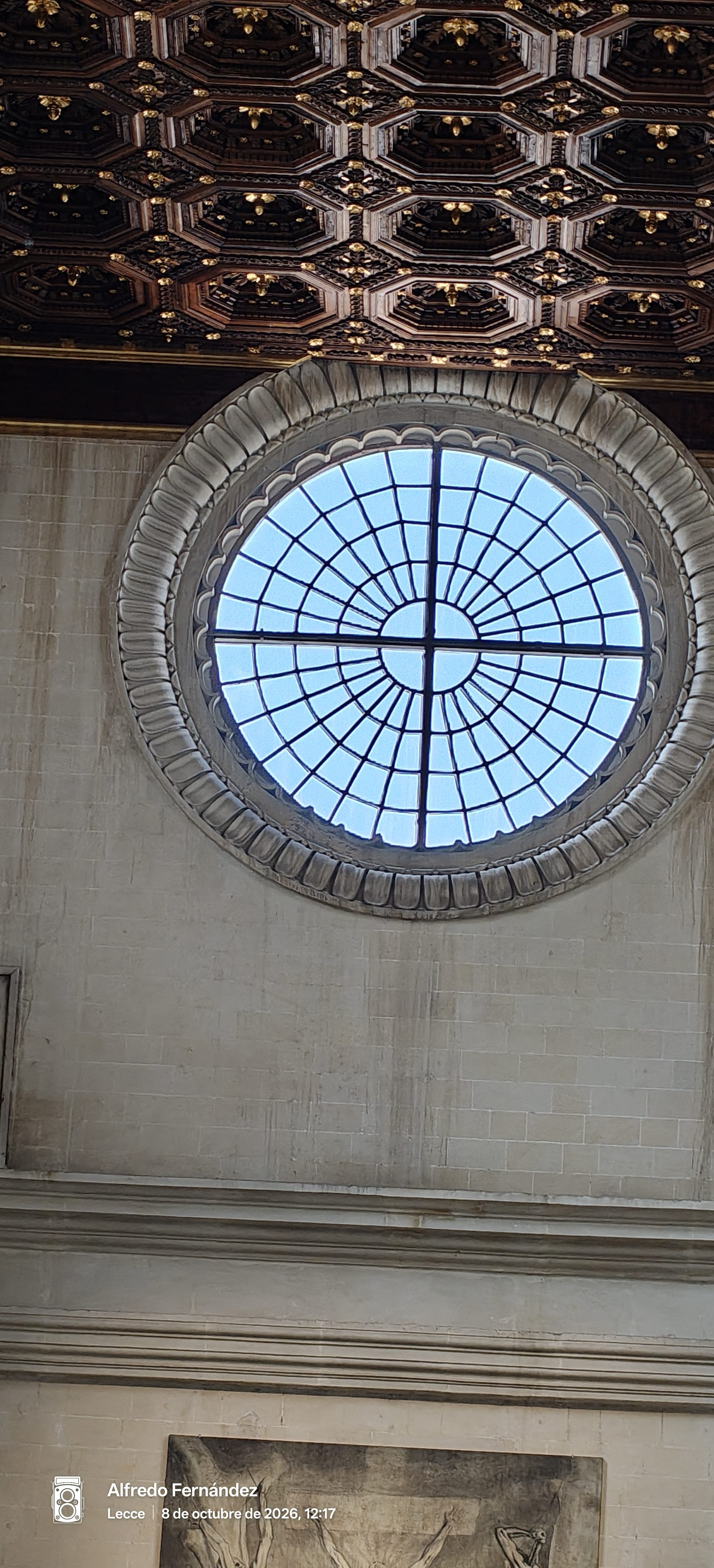 Ventana circular de piedra bajo un elaborado artesonado de madera tallada y dorada.