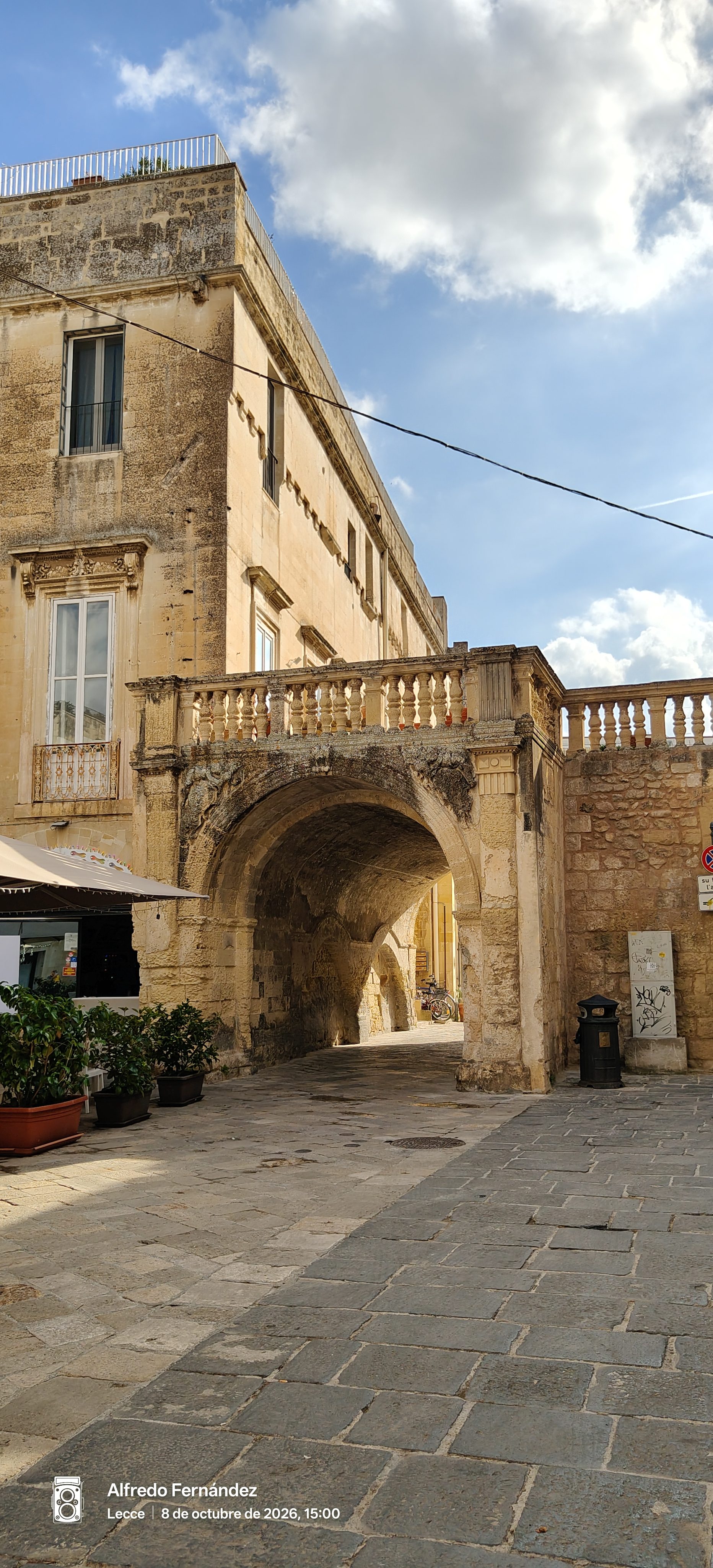Arco de piedra histórica y terraza con balaustrada en una calle empedrada soleada.
