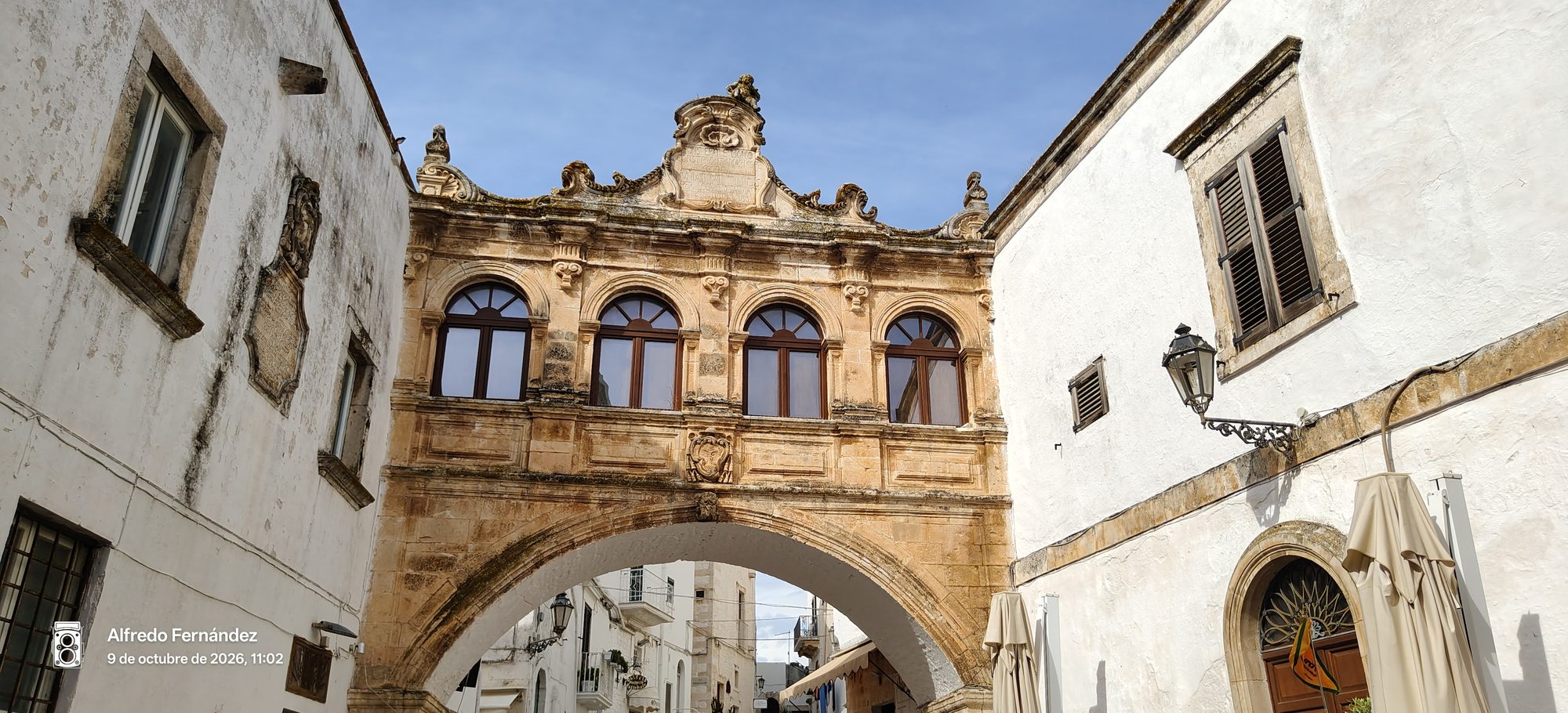 Arco de piedra ornamentado conectando edificios blancos bajo el cielo azul en Ostuni.
