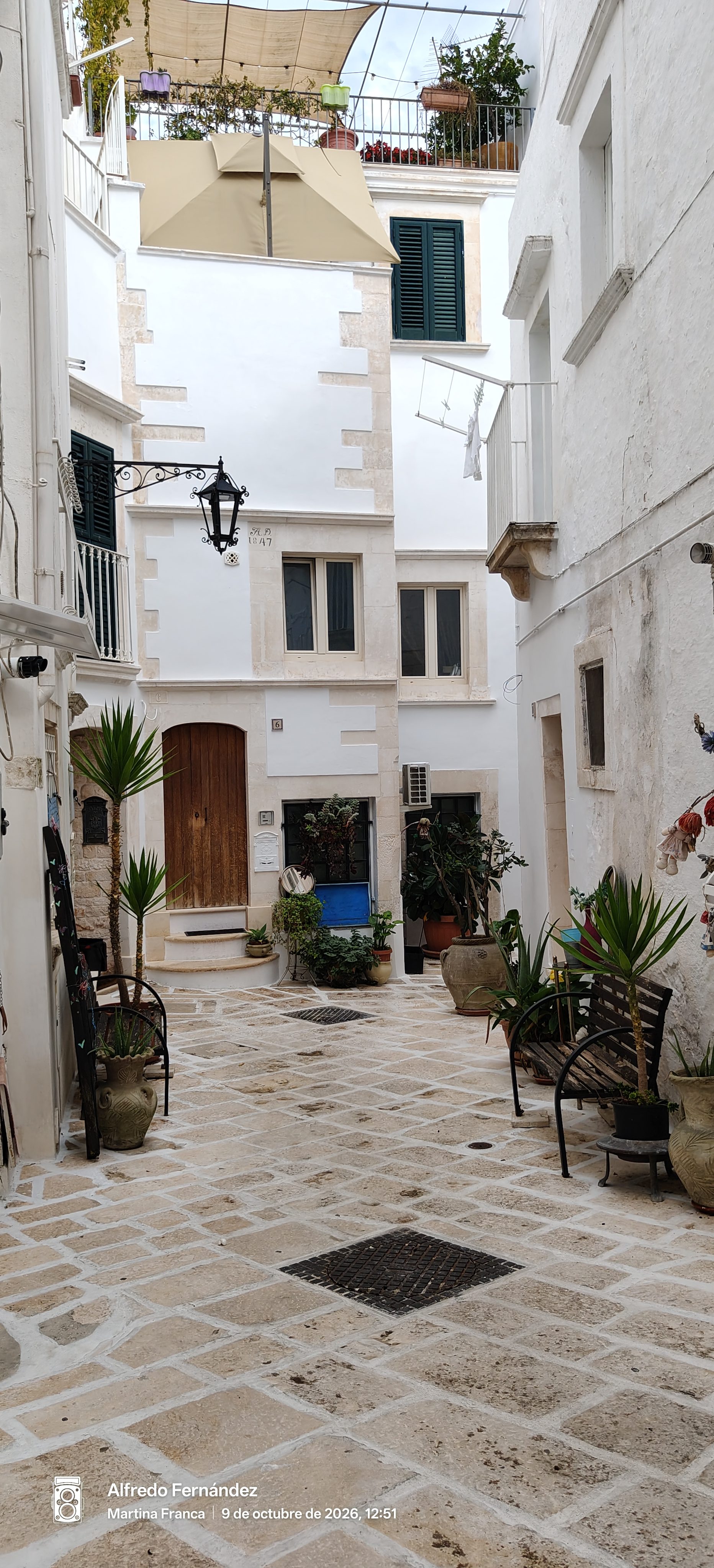 Callejón empedrado con fachadas blancas, plantas en macetas y bancos en Martina Franca.