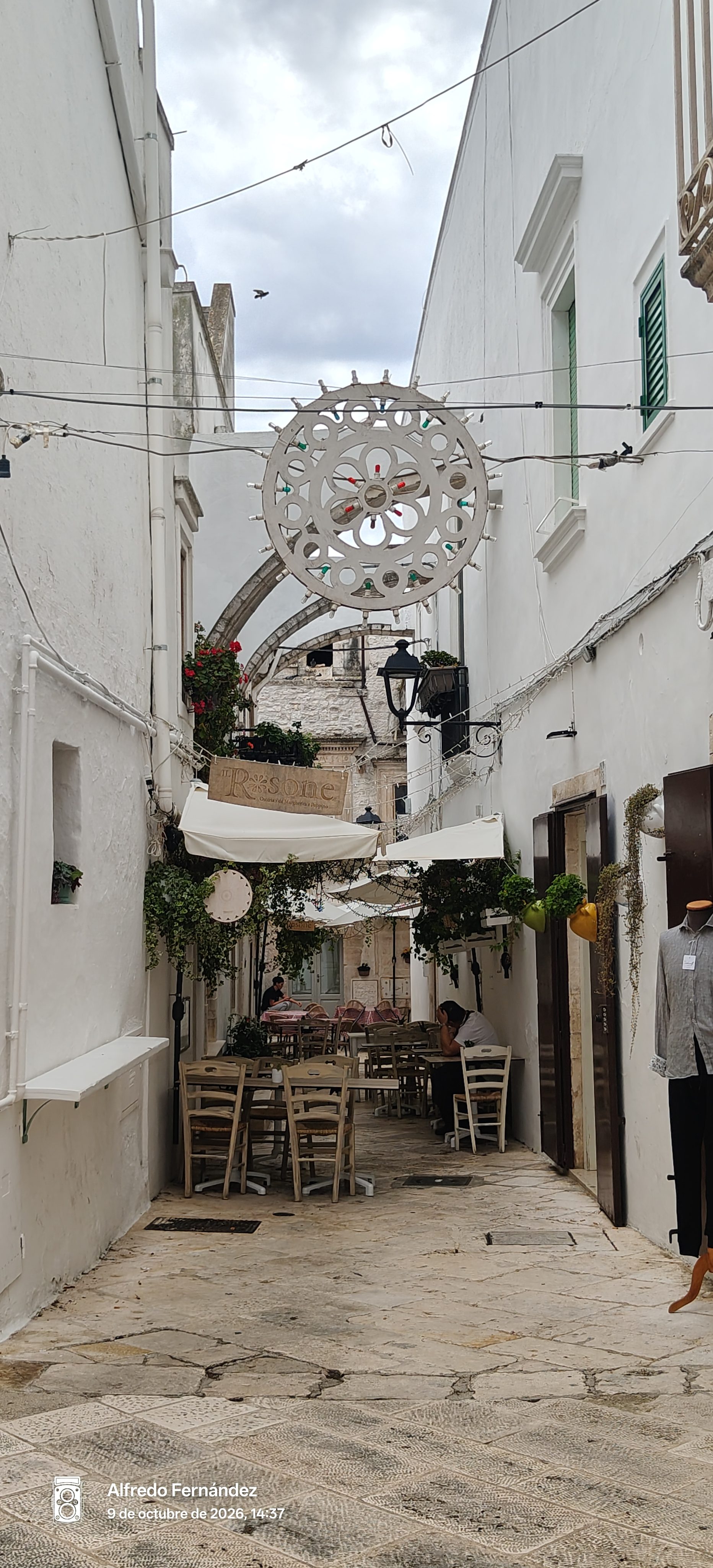 Callejuela blanca empedrada con mesas al aire libre y una roseta decorativa en Locorotondo.