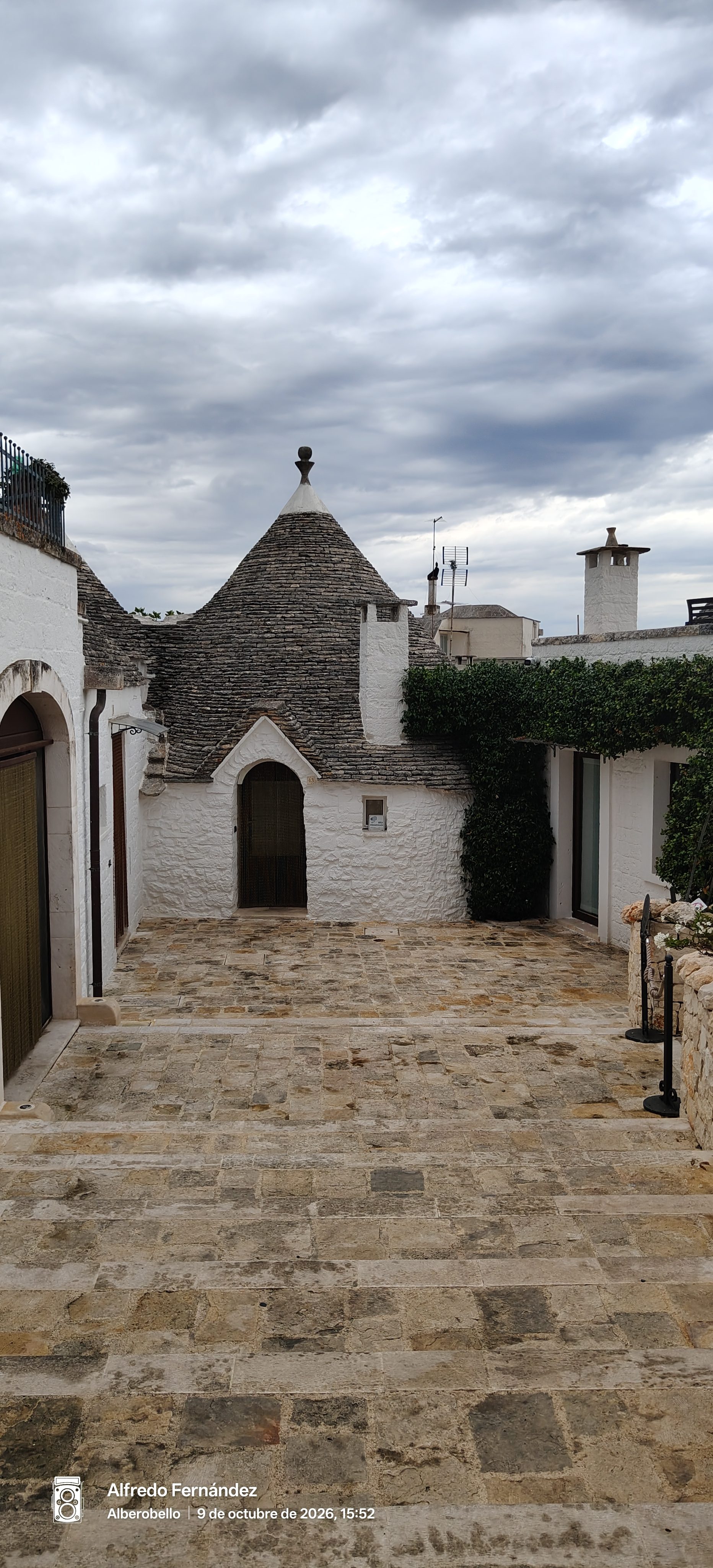 Trullo tradicional de piedra blanca con tejado cónico y patio empedrado bajo cielo nublado.