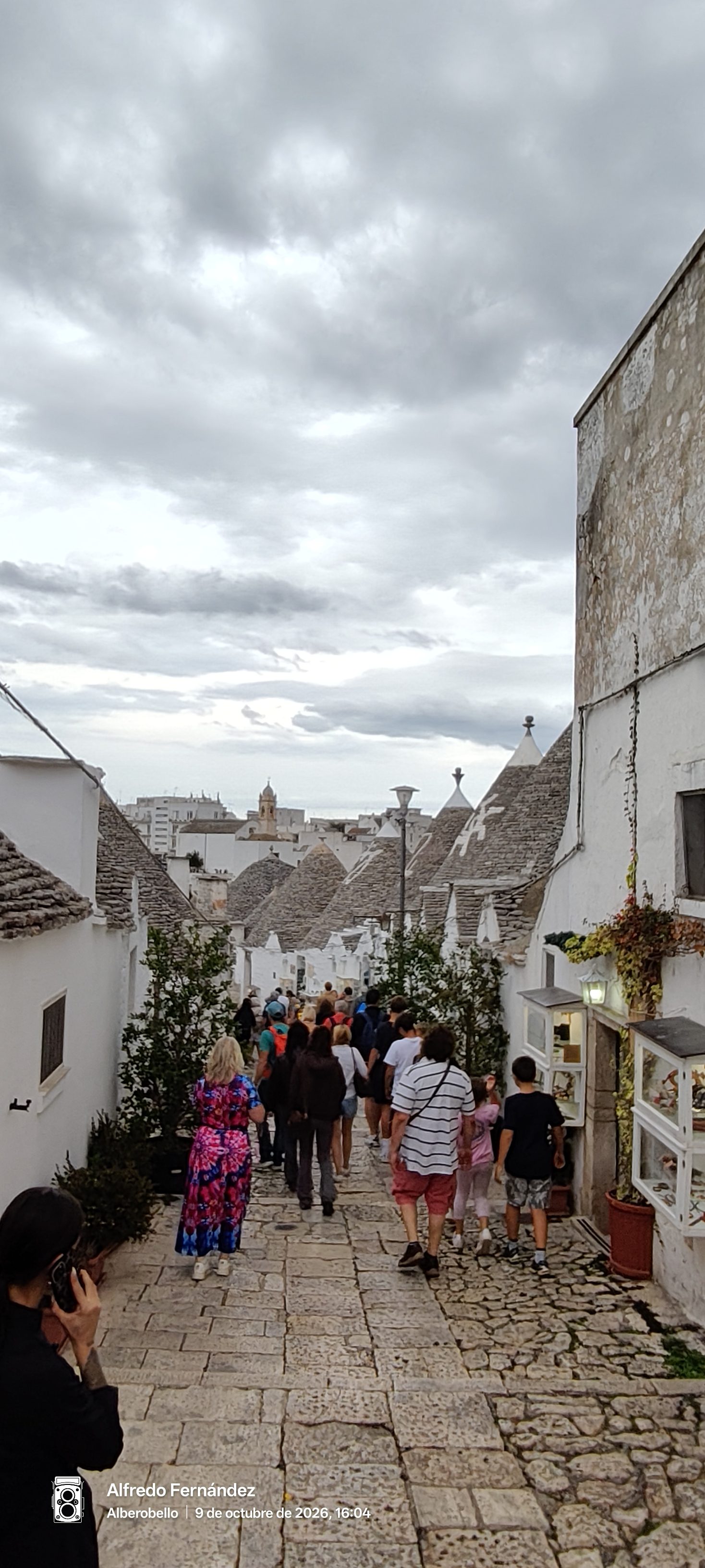 Calle empedrada descendente flanqueada por casas trulli tradicionales bajo un cielo nublado.