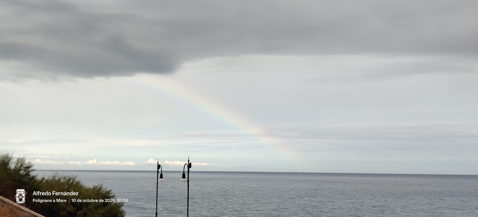 Arcoíris tenue sobre el mar bajo un cielo nublado con farolas al frente.