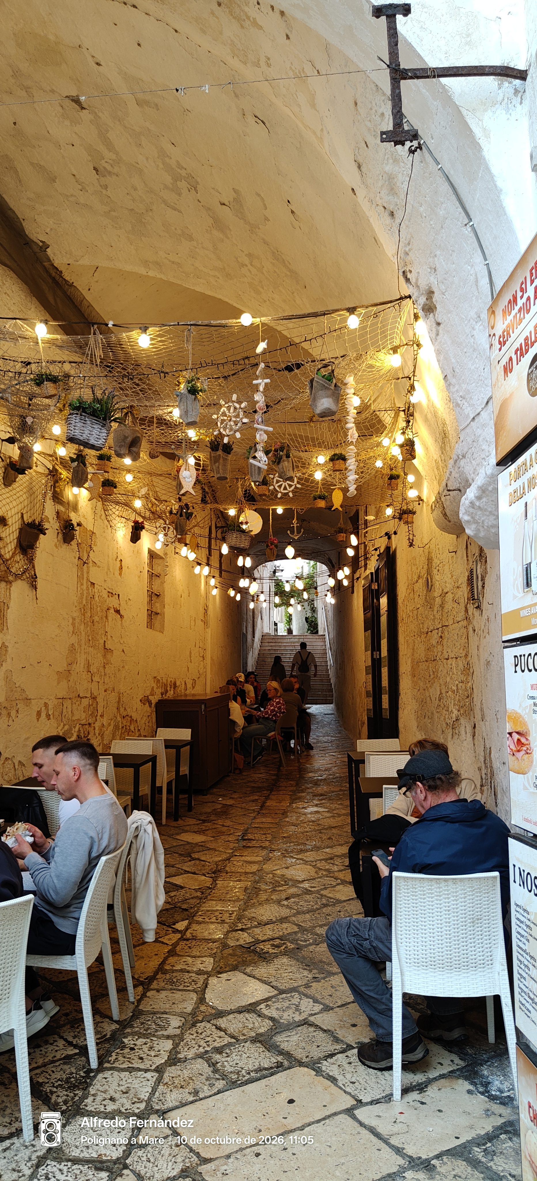 Callejón abovedado decorado con luces y redes marinas entre mesas de un restaurante.
