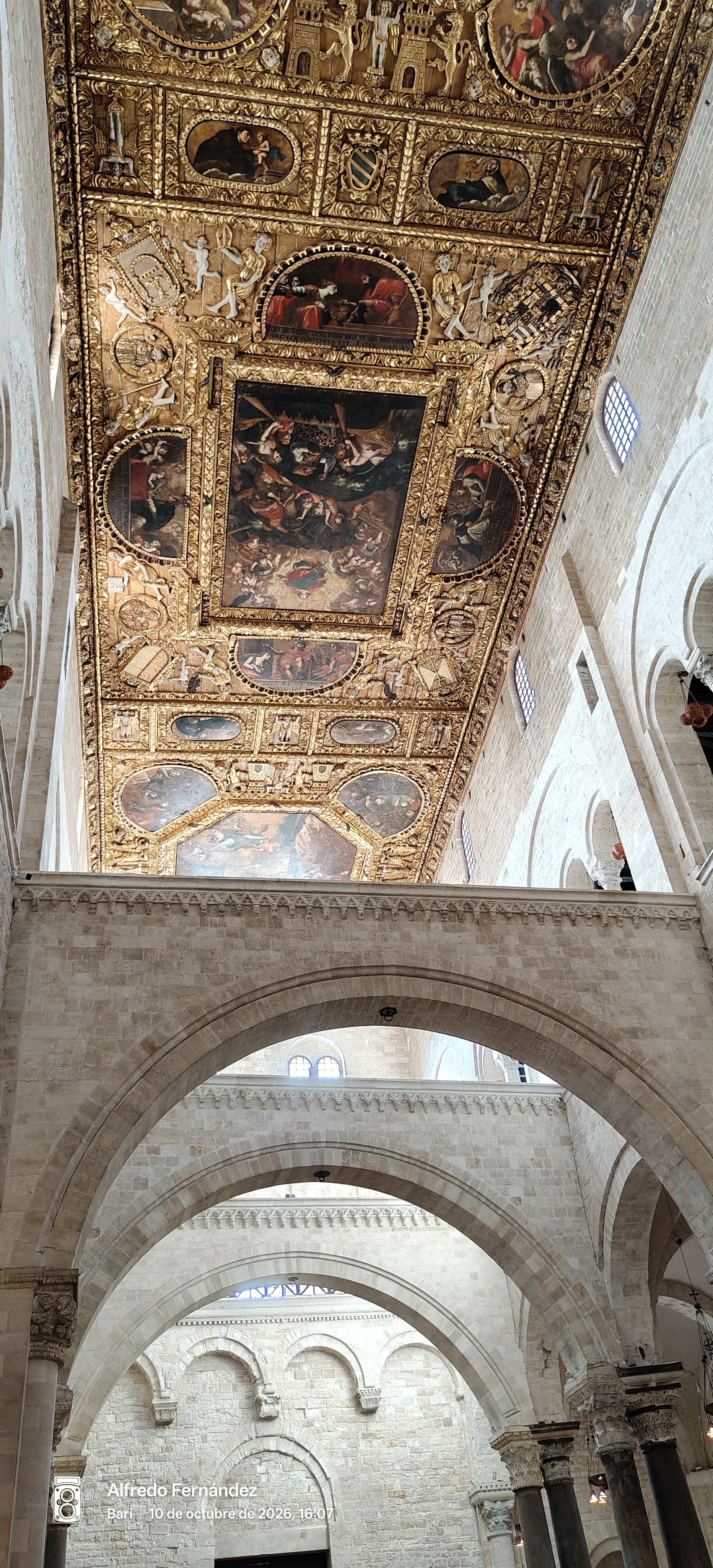 Techo artesonado dorado con pinturas religiosas sobre arcos románicos de piedra en una basílica.