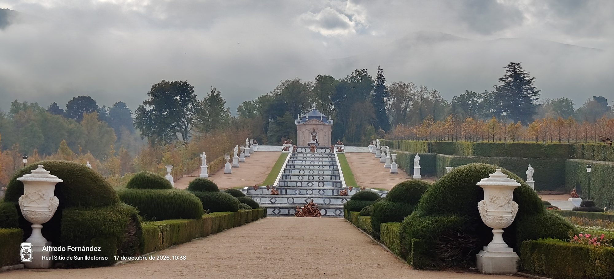 Cascada monumental y jardines formales con estatuas y setos bajo un cielo nublado.
