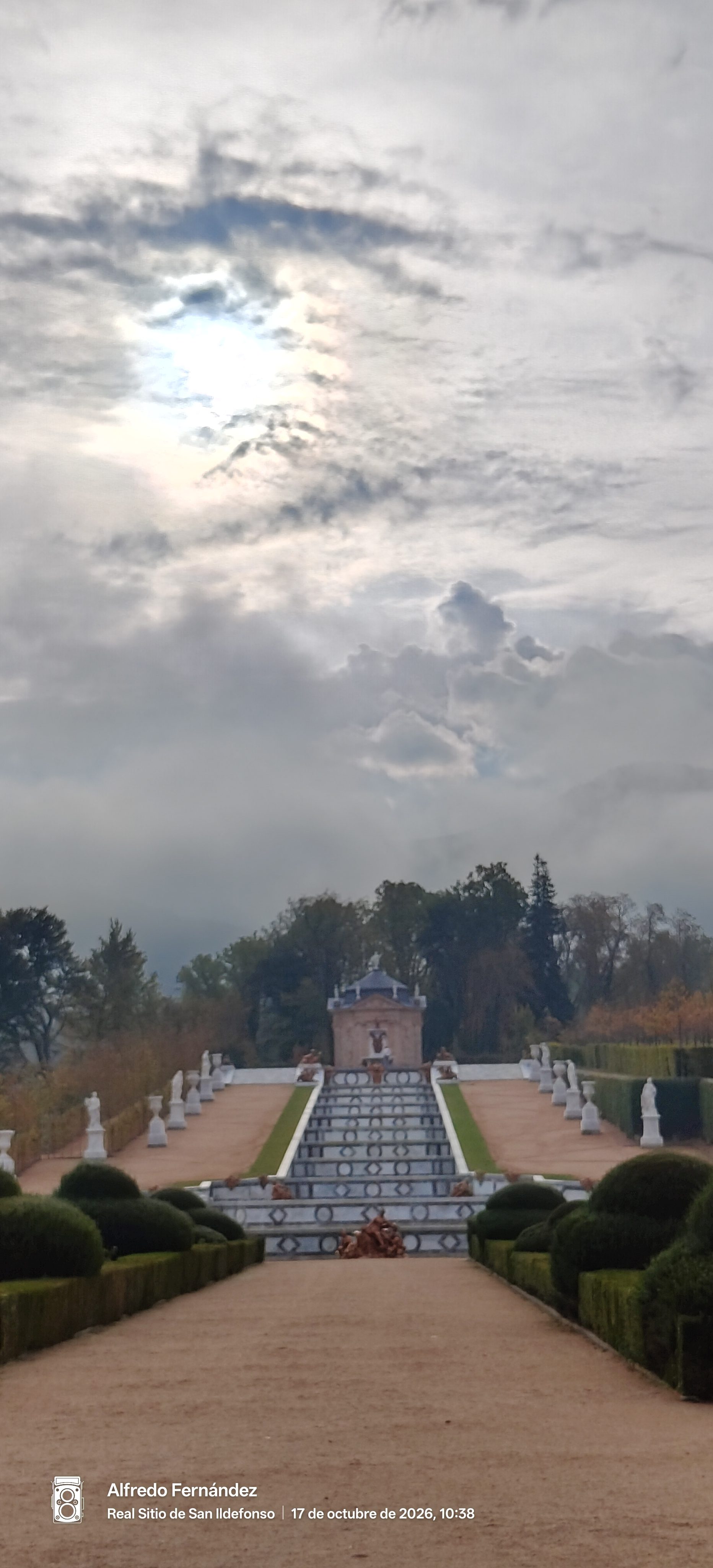 Cascada monumental y estatuas en jardines históricos bajo un cielo nublado con sol velado.