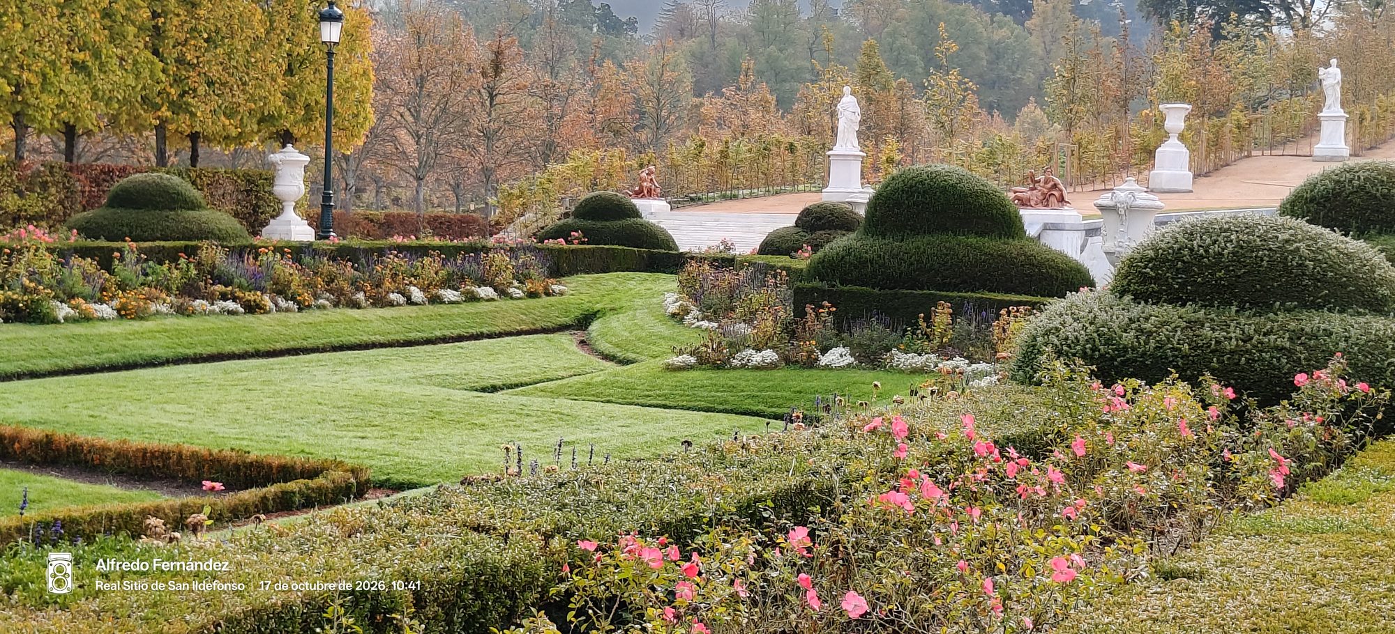 Jardines geométricos con setos recortados, flores y estatuas clásicas rodeados de árboles otoñales.