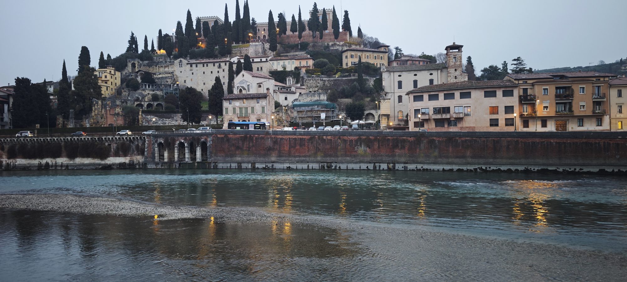 Colina histórica con cipreses y edificaciones antiguas reflejadas en el río Adigio de Verona.