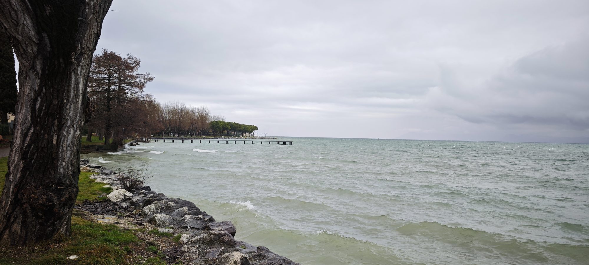Orilla rocosa del lago de Garda bajo un cielo nublado invernal con muelle al fondo.