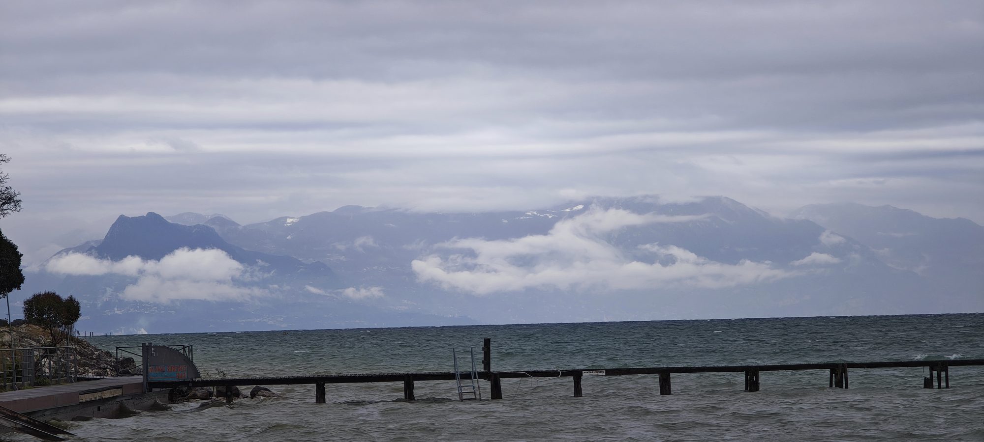 Embarcadero de madera en el lago con montañas entre nubes bajo un cielo nublado.