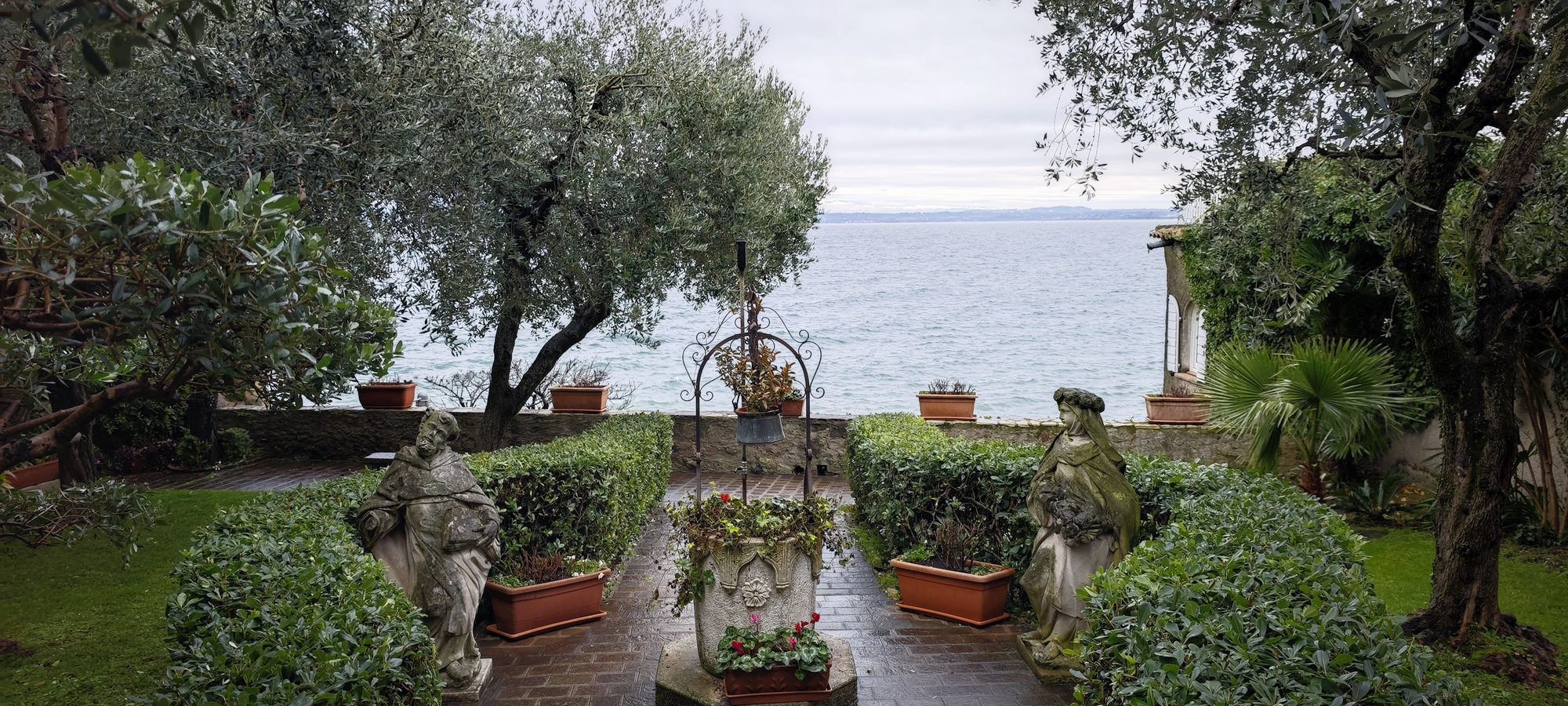 Jardín junto al lago de Garda con estatuas de piedra, olivos y un pozo.