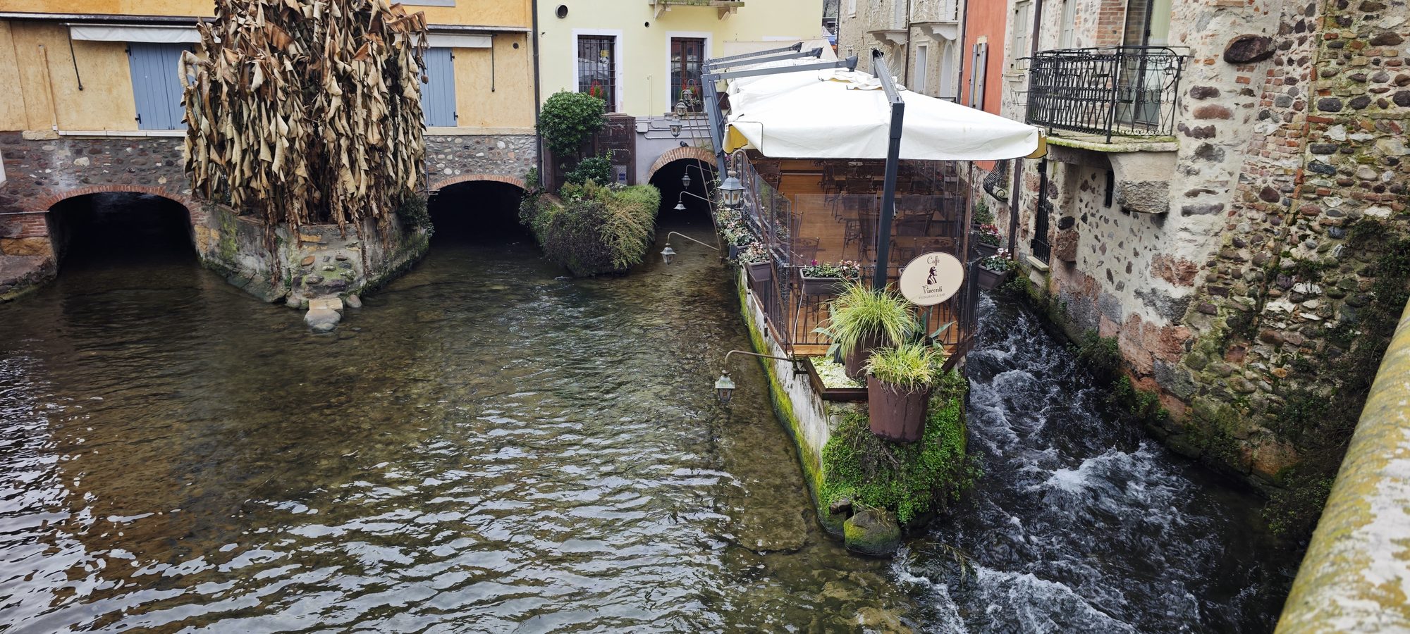 Canal de agua cristalina fluyendo entre antiguos edificios de piedra y una terraza rústica.