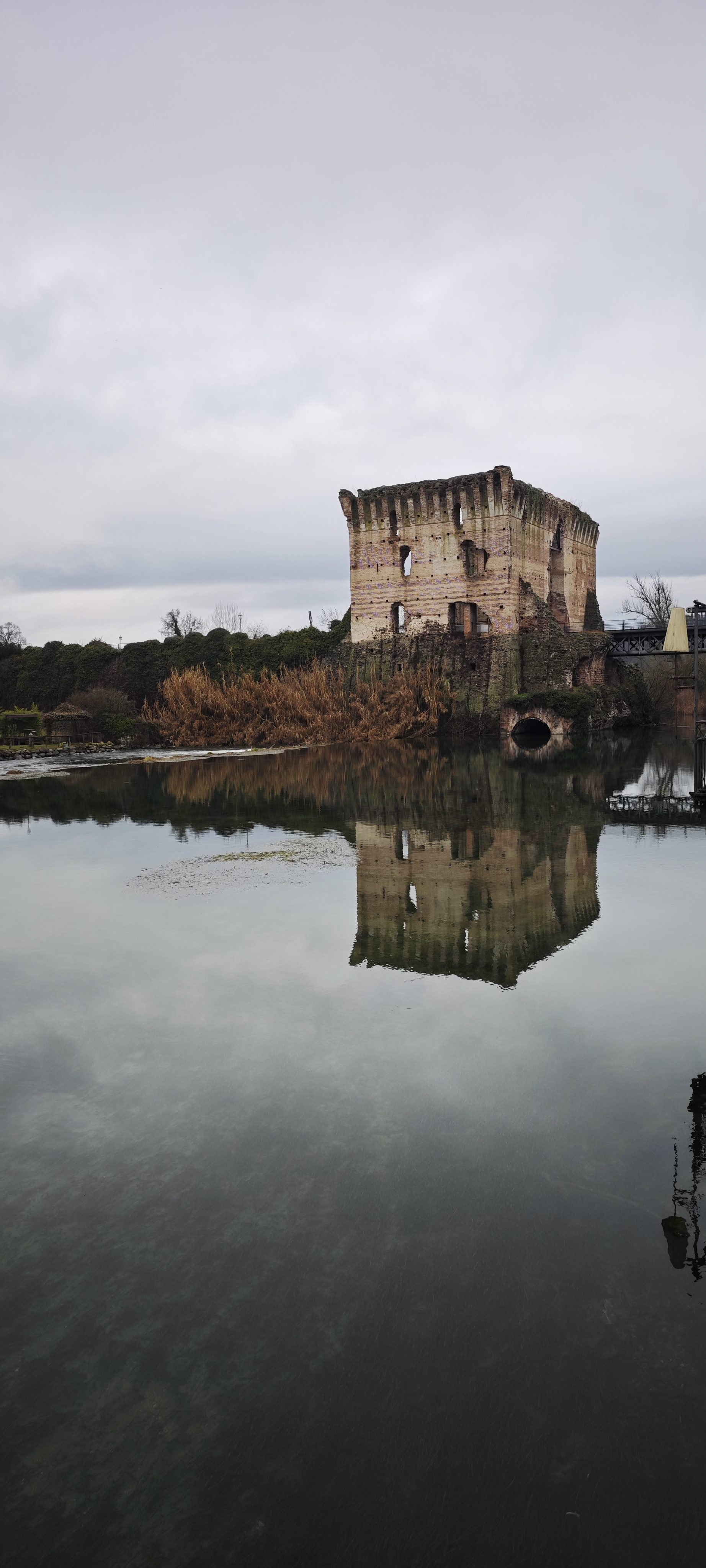 Torre medieval en ruinas reflejada en el río Mincio bajo un cielo nublado.