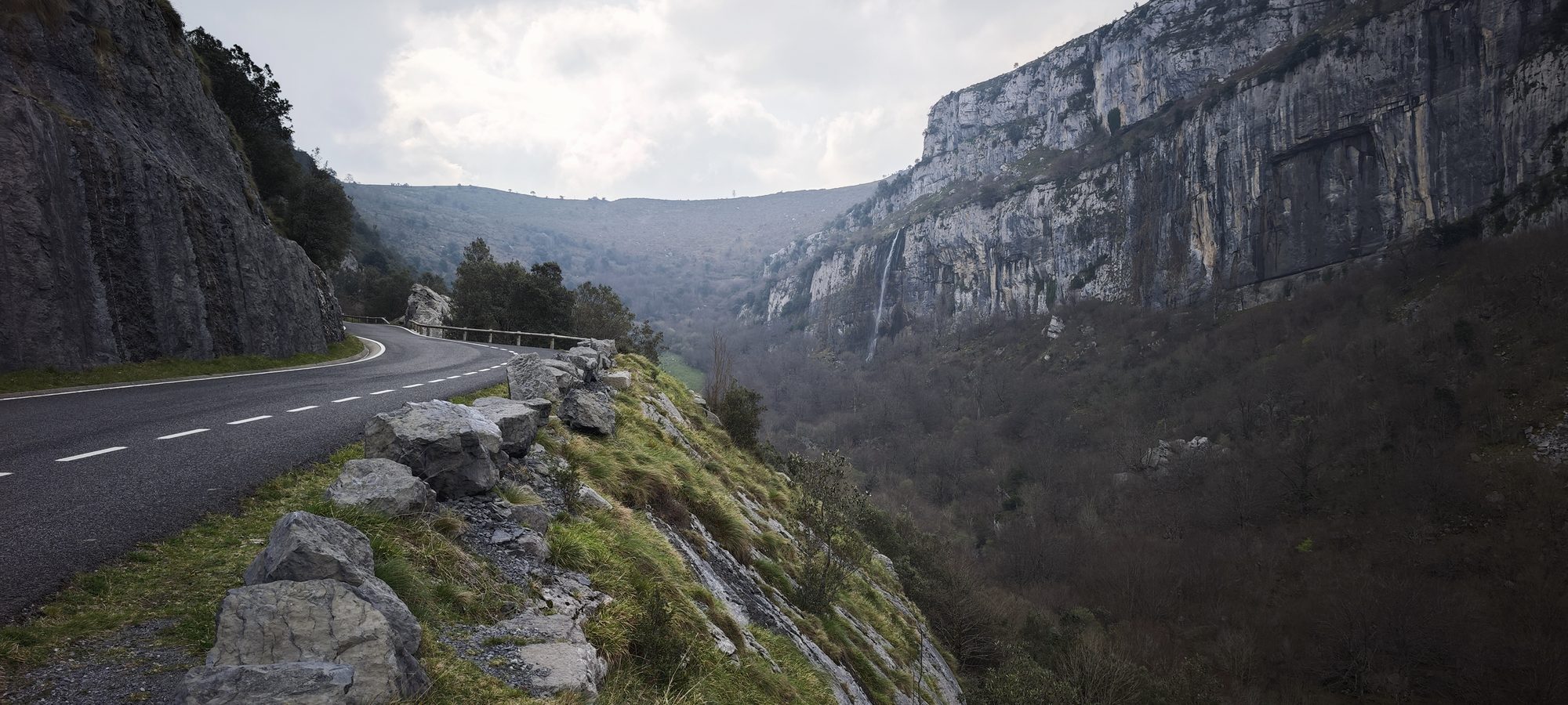 Carretera de montaña bordeando un desfiladero rocoso con una cascada lejana en el valle.