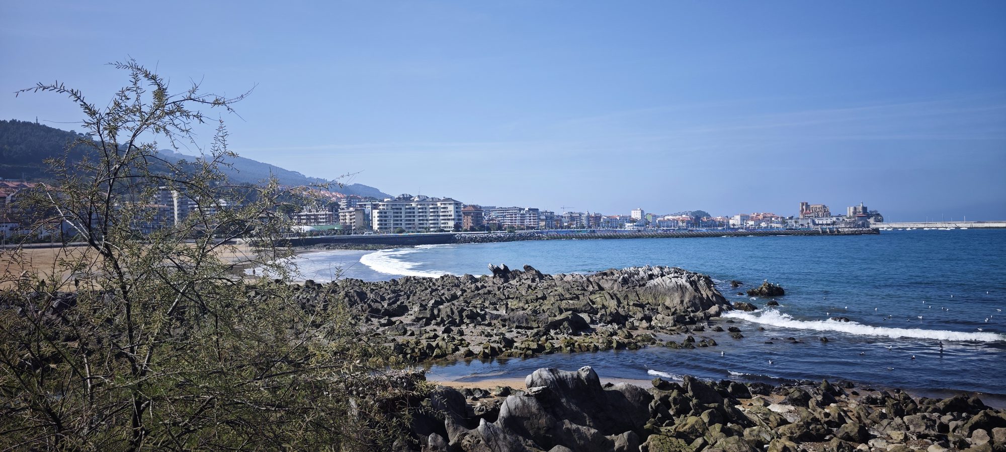 Vista de costa rocosa, playa y ciudad costera junto al mar bajo cielo claro.