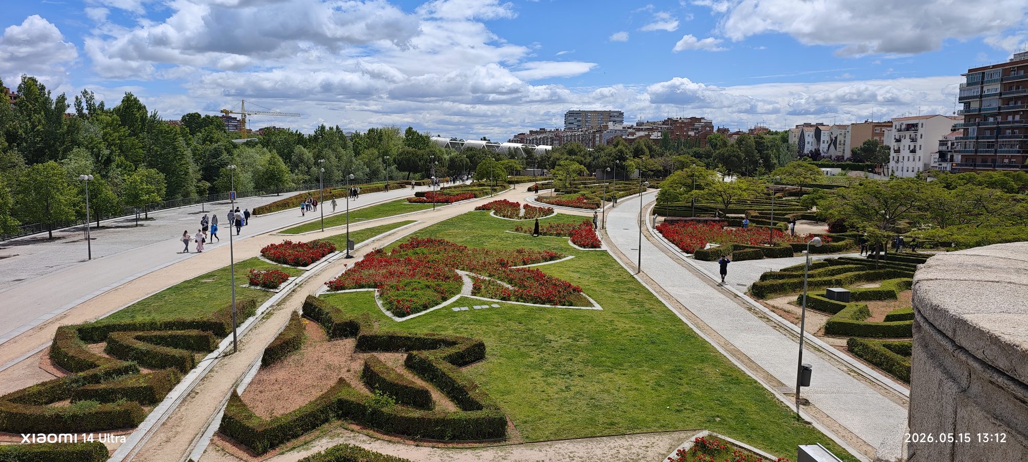 Vista panorámica de un parque urbano con jardines geométricos, caminos peatonales y edificios al fondo.