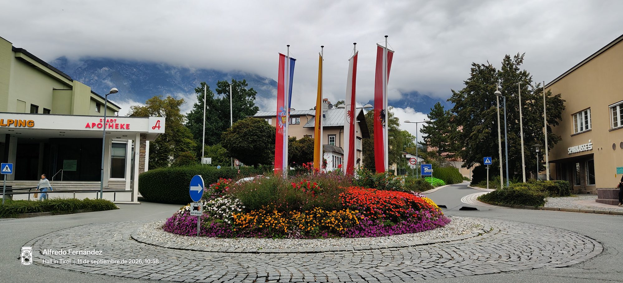 Rotonda decorada con flores coloridas y banderas regionales con montañas alpinas de fondo.