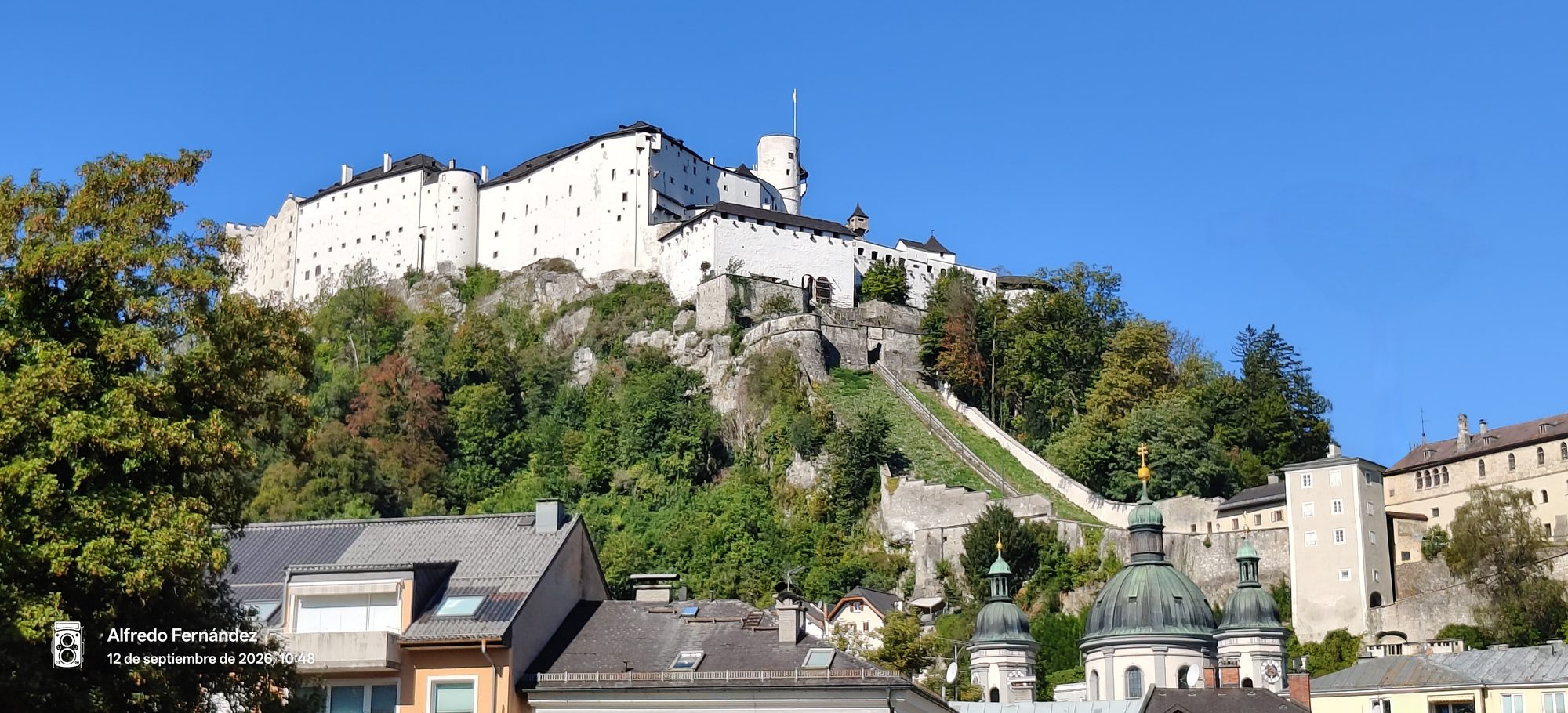 Fortaleza de Hohensalzburg sobre colina boscosa con tejados y cúpulas en primer plano.