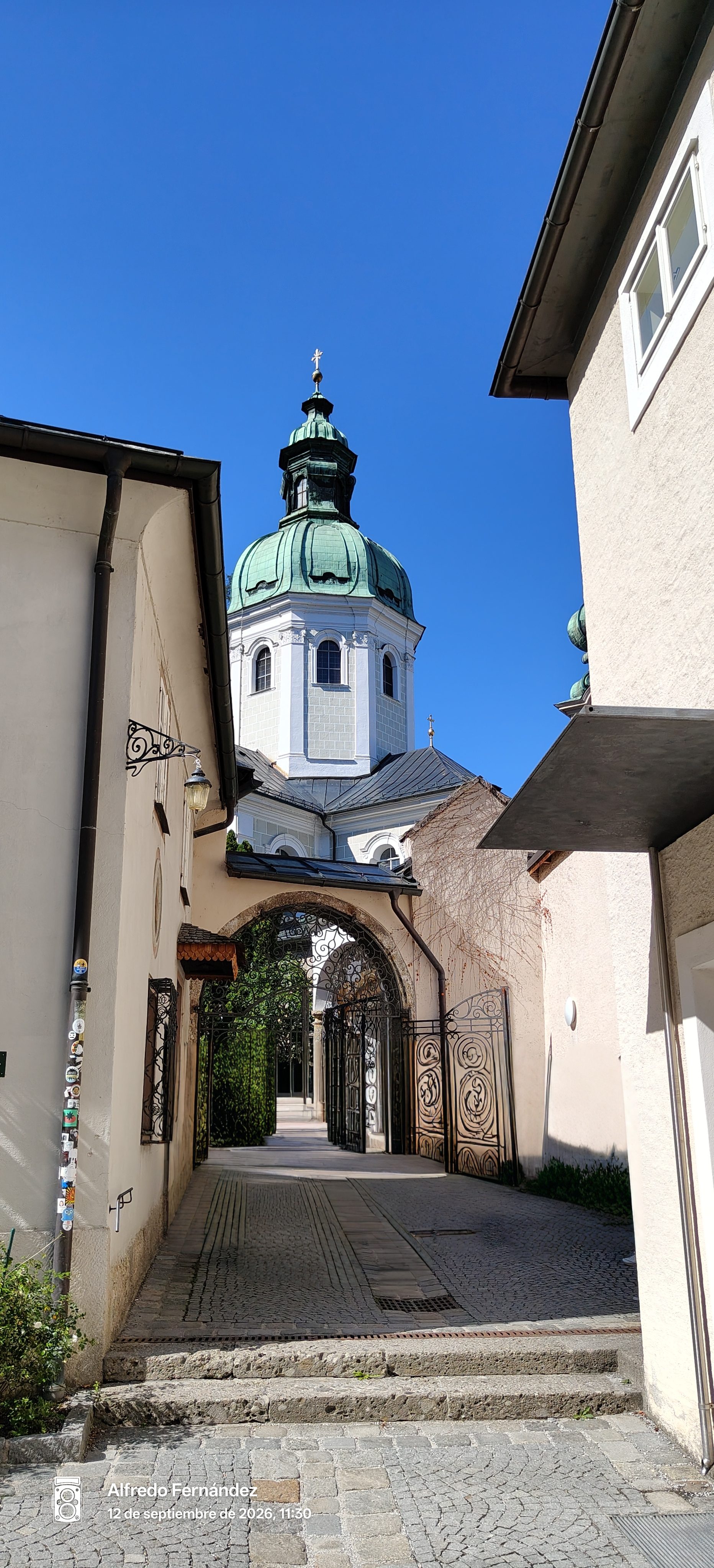 Callejón adoquinado con cancela de hierro forjado hacia la cúpula de una iglesia barroca.