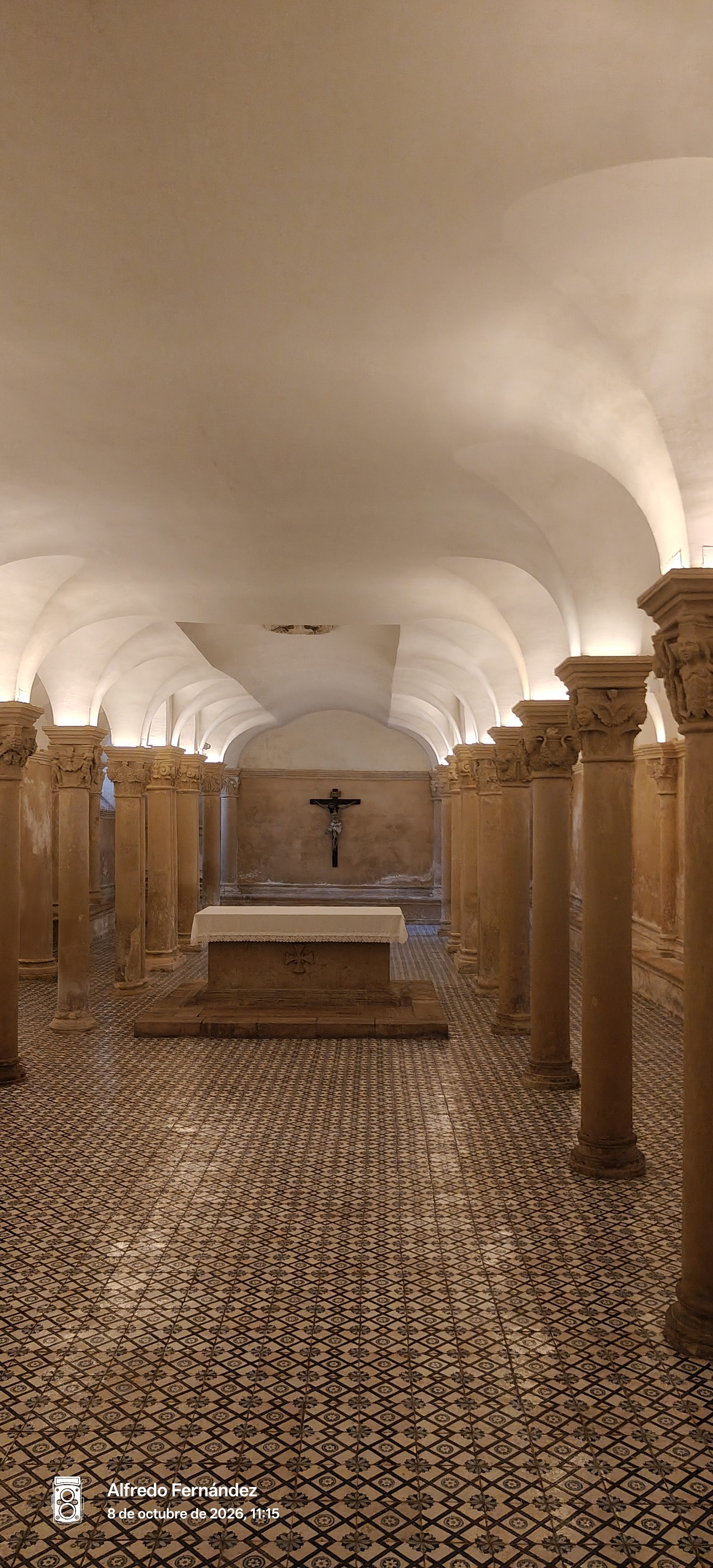 Cripta subterránea con columnas de piedra, suelo de mosaico y un altar con crucifijo.